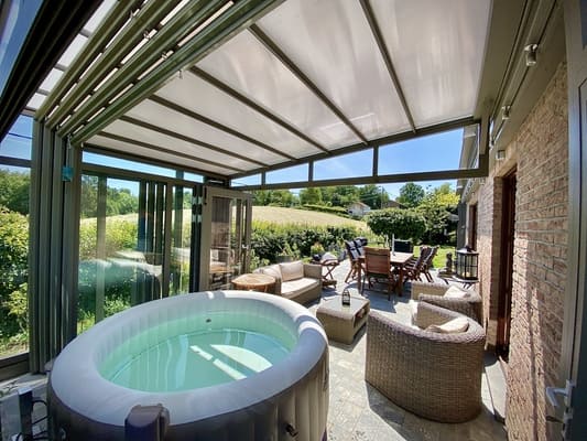 Une véranda rétractable abritant le spa et ouverte sur la terrasse avec des fauteuils, des chaises et une table