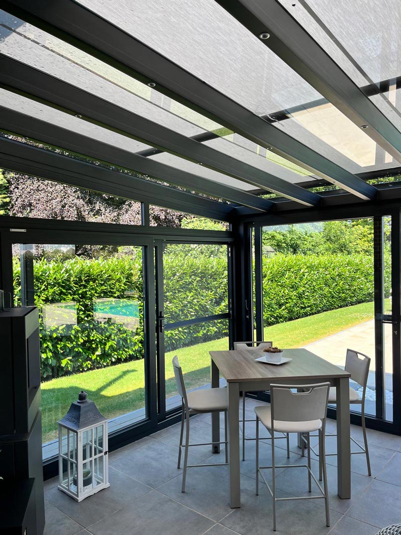 Coin repas sous une véranda moderne avec toit en verre et structure en aluminium noir, équipée d'une table haute en bois et de chaises blanches.