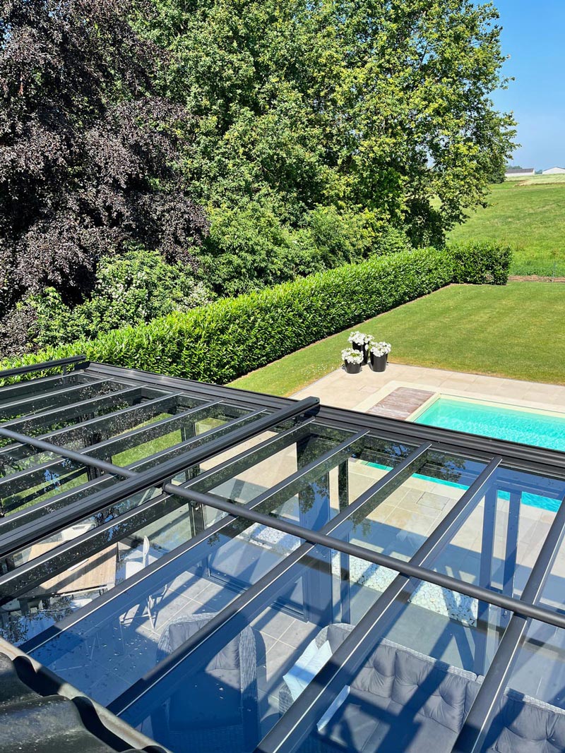 Vue aérienne d'une véranda avec toit en verre, montrant l'intérieur aménagé avec des meubles en rotin et l'extérieur avec une piscine et un jardin verdoyant.