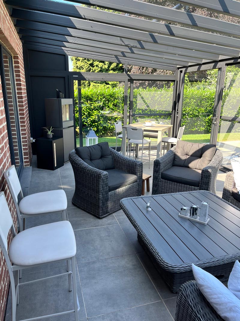 Espace de vie extérieur couvert avec des fauteuils en rotin gris, une table basse en bois, et un coin repas avec des chaises blanches, sous une véranda en aluminium gris.